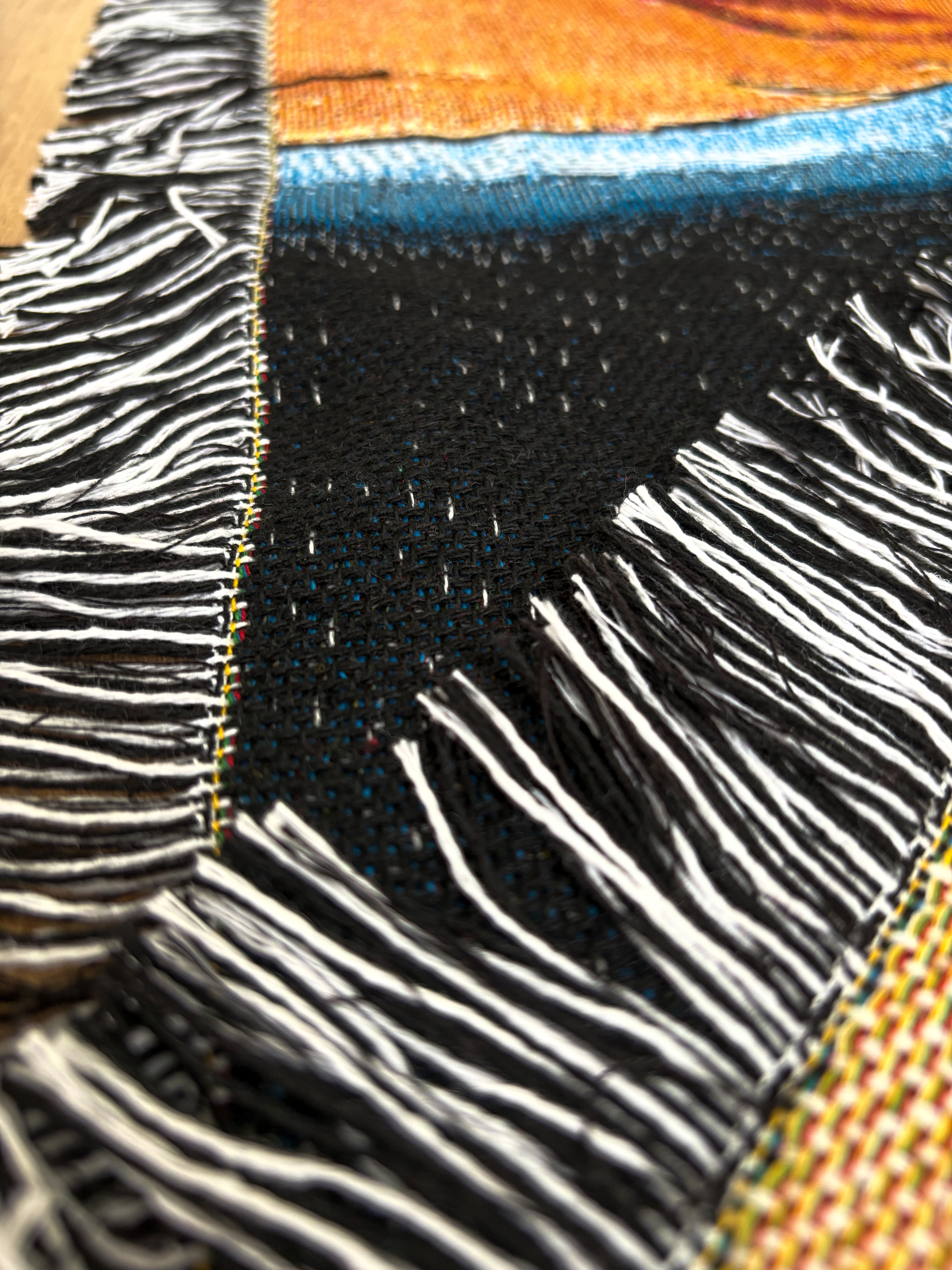 Close-up of a woven textile with black and white fringe, a dark central section, and visible orange, blue, and yellow woven areas.